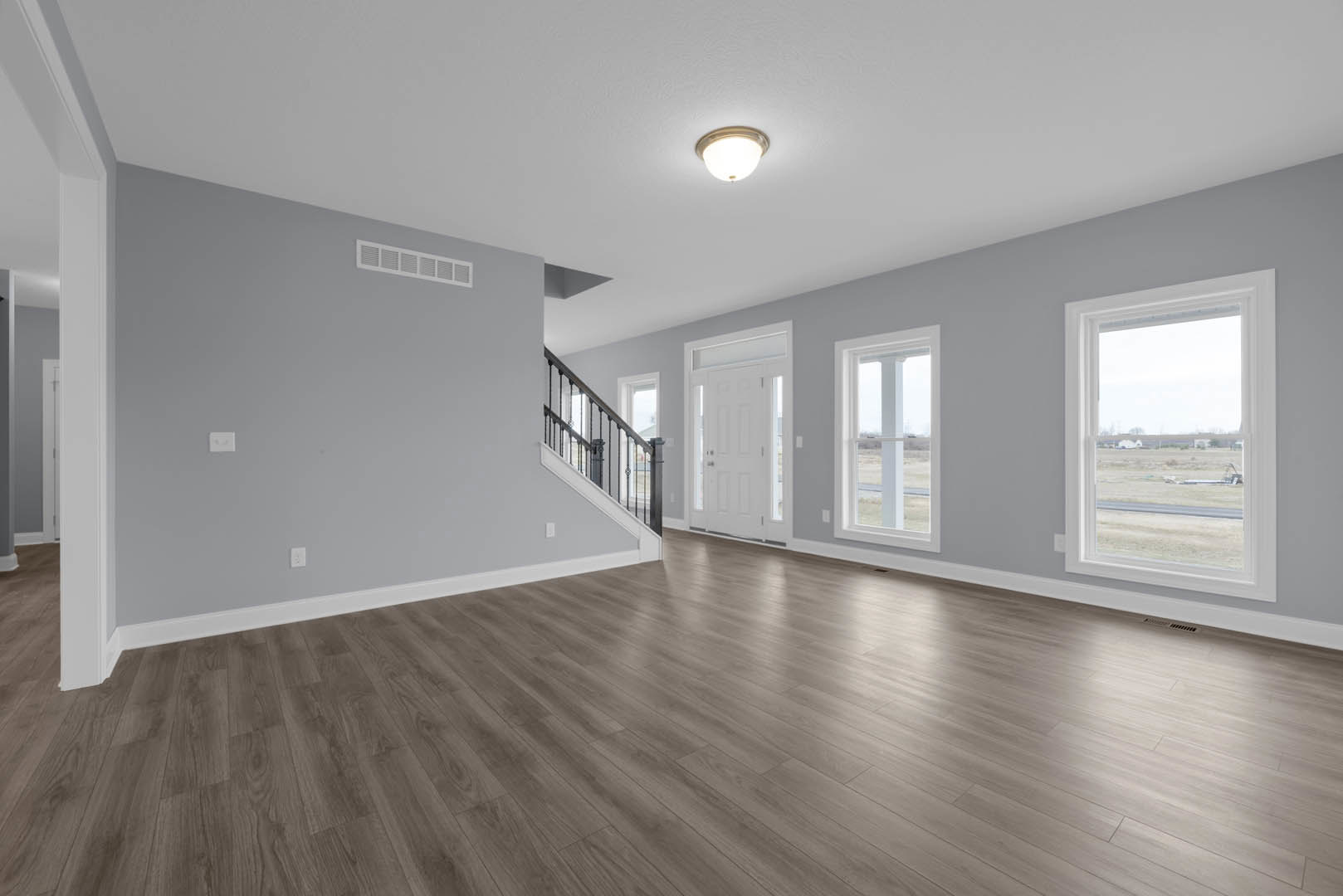 Wood flooring in a bright room with white door, window overlooking open field, staircase detail, ceiling light fixture, and plaster walls with molding.