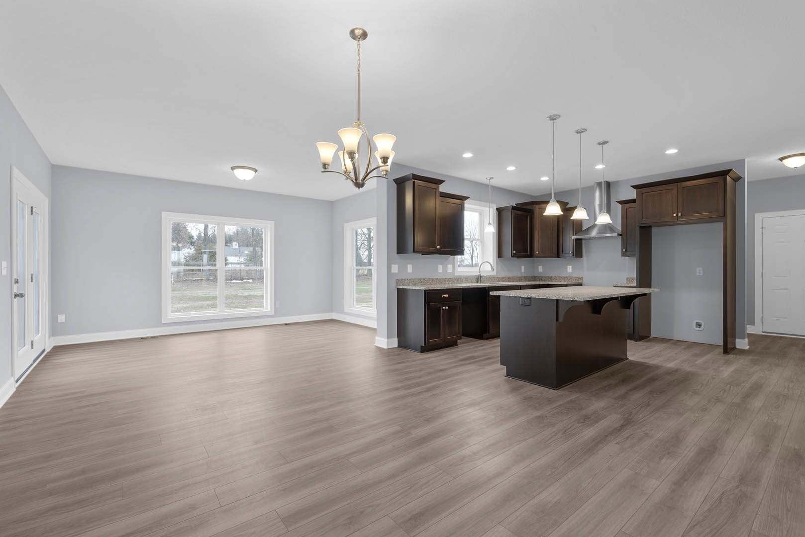 Open kitchen and dining area featuring wood flooring, marble-topped island, white cabinetry, rectangular-patterned door, large window overlooking yard, and modern chandelier.