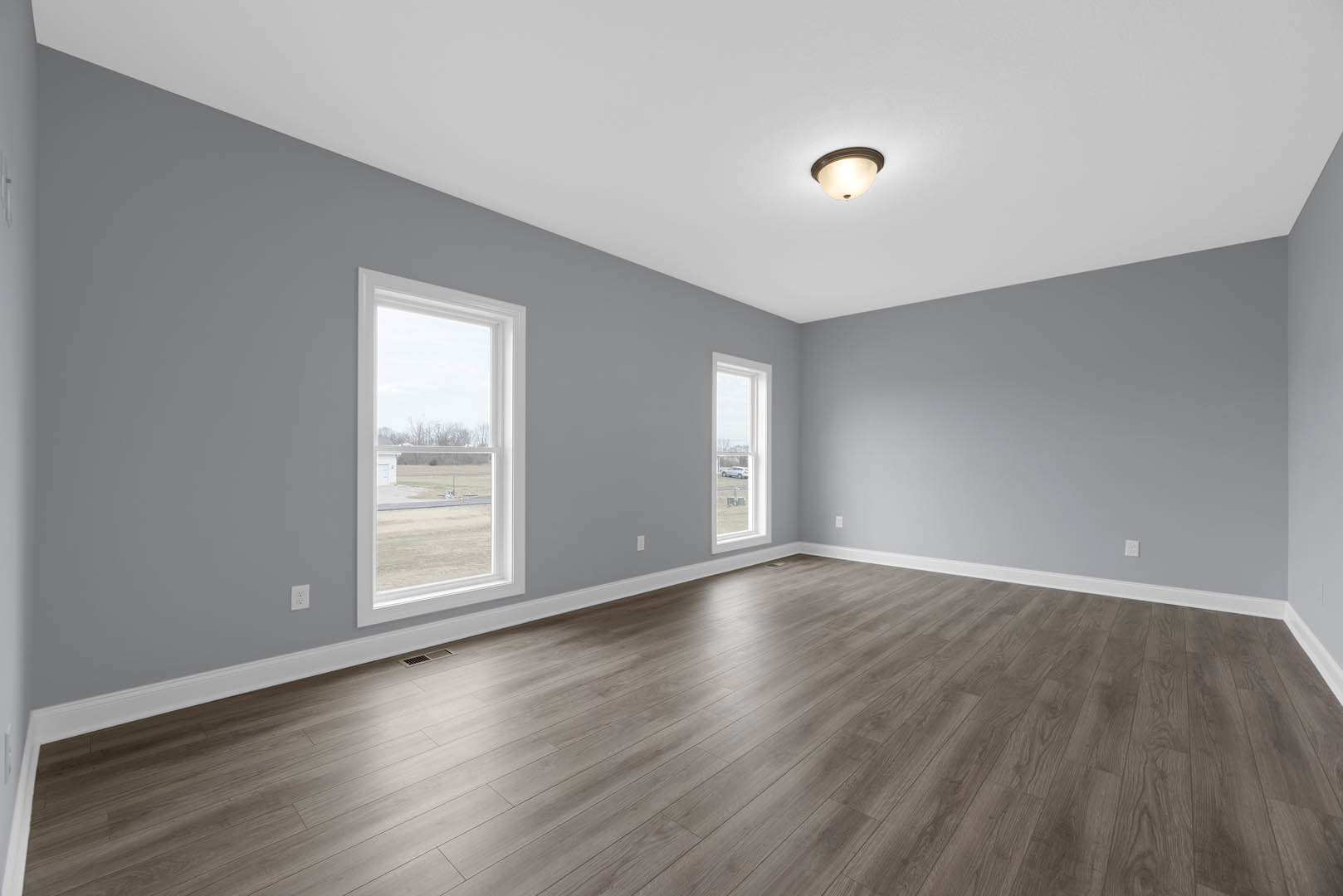 Open room with light wood flooring, multiple large windows showing views of a field, road, and parked car, white plaster walls, and modern ceiling light fixture