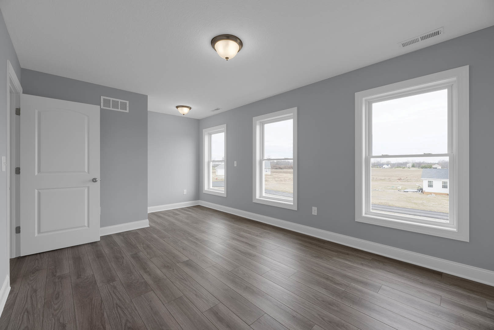 Bright room with wood flooring, white walls, ceiling light fixture, white door with silver knob, and windows offering views of a field and neighboring house