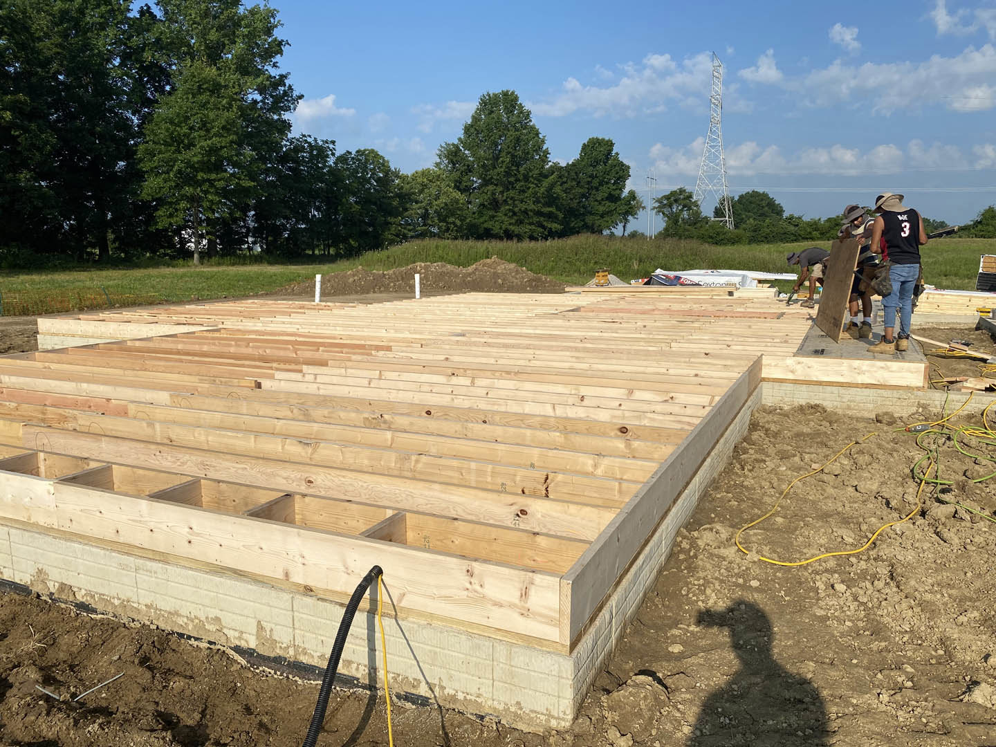 Exposed wooden framing of a house under construction with a person in black shirt and blue jeans standing nearby, metal tower in background, yellow and green hose on dirt, and
