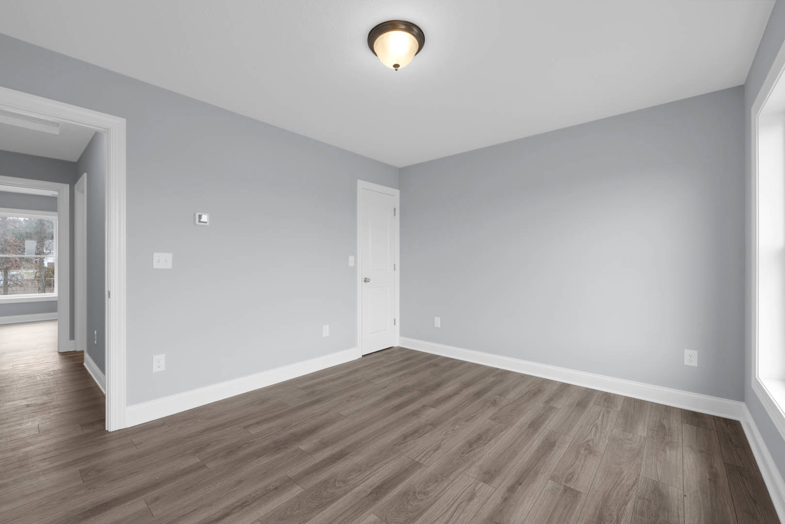 White door with silver knob, wood flooring with white base trim, ceiling light fixture, plaster walls, window partially visible