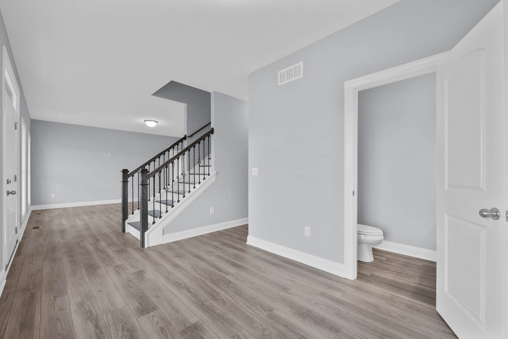 Hardwood floor entryway with white-painted staircase, paneled door, white wall vent, and partial view of a closed toilet in adjacent room