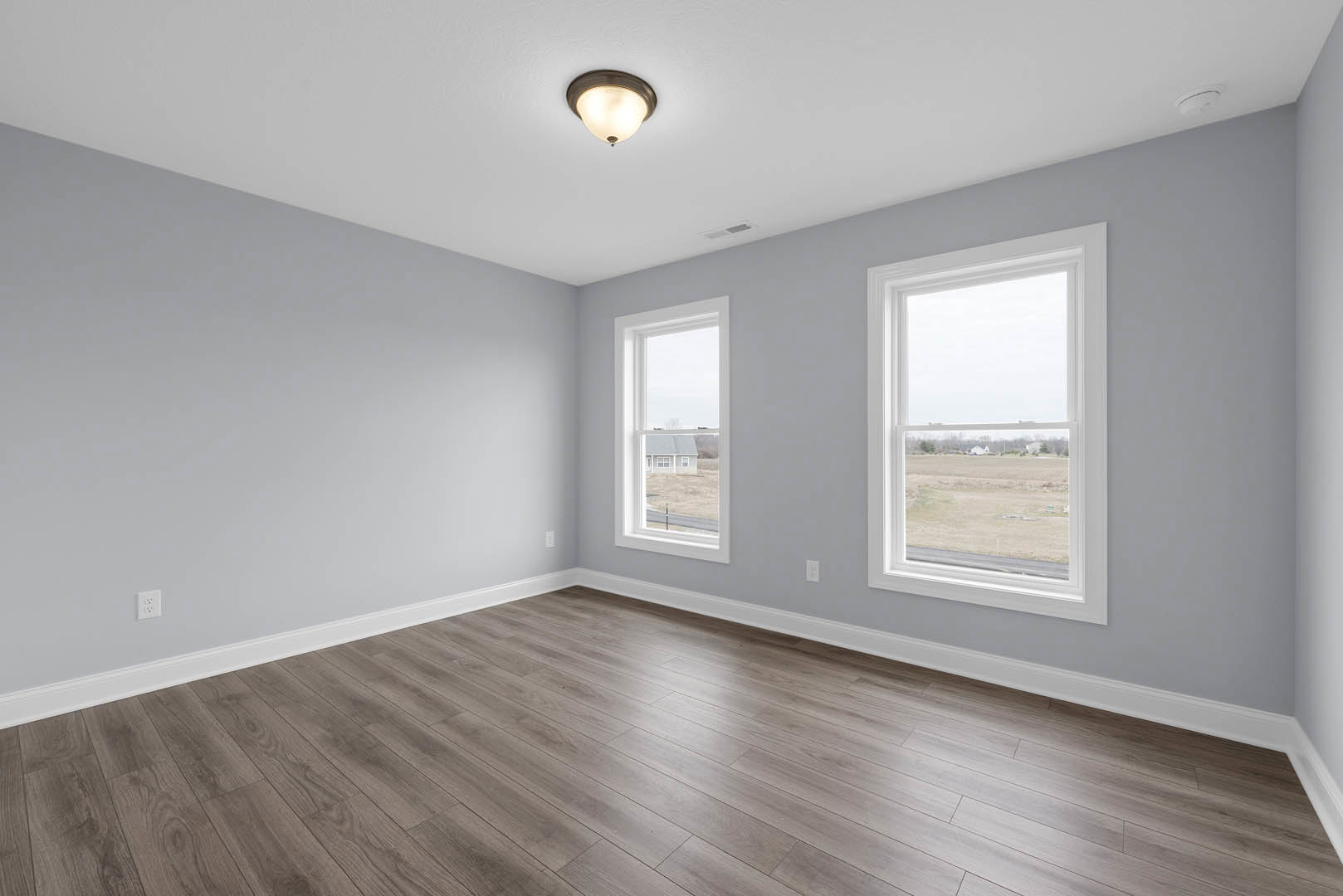 Spacious room with light wood flooring, large windows overlooking a field, white plaster walls, ceiling-mounted circular light fixture, and decorative molding.