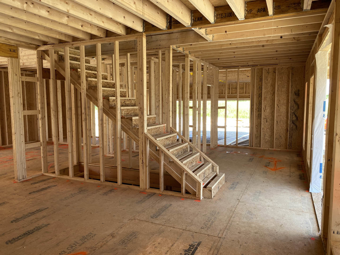 Exposed wood framing with unfinished beams, partially constructed wooden staircase, stone accent wall with white trim, subfloor and insulation visible in custom home interior