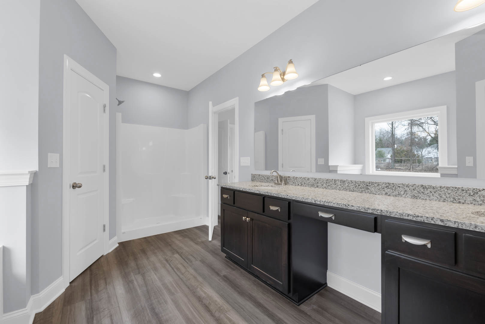 Bathroom featuring marble countertop, wood flooring, white door, three-light fixture, window with view of trees, and white rectangular wall light