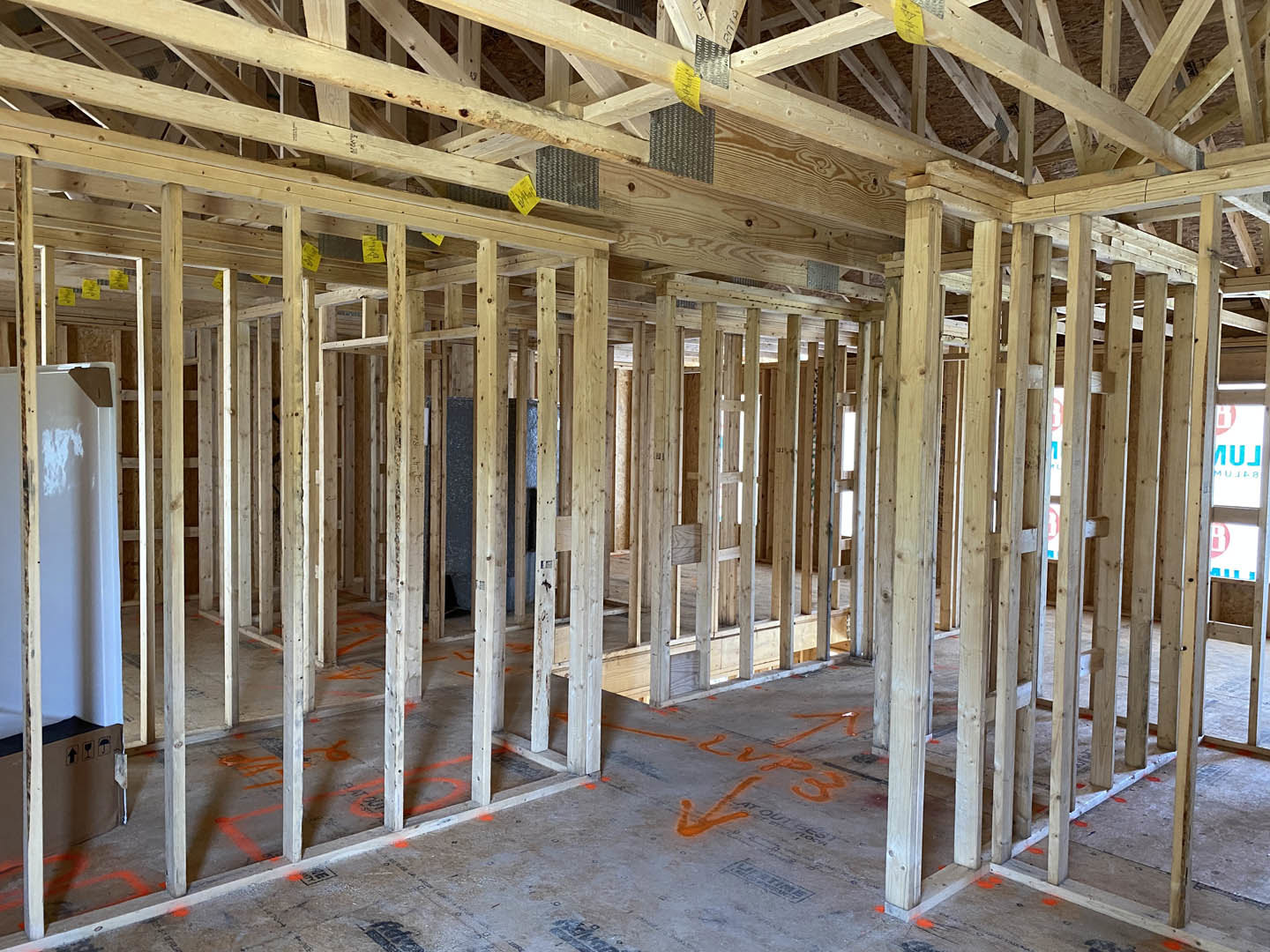 Wooden house frame under construction with exposed beams, roof trusses, and orange paint on the subfloor; white pole and blue wall visible in background.