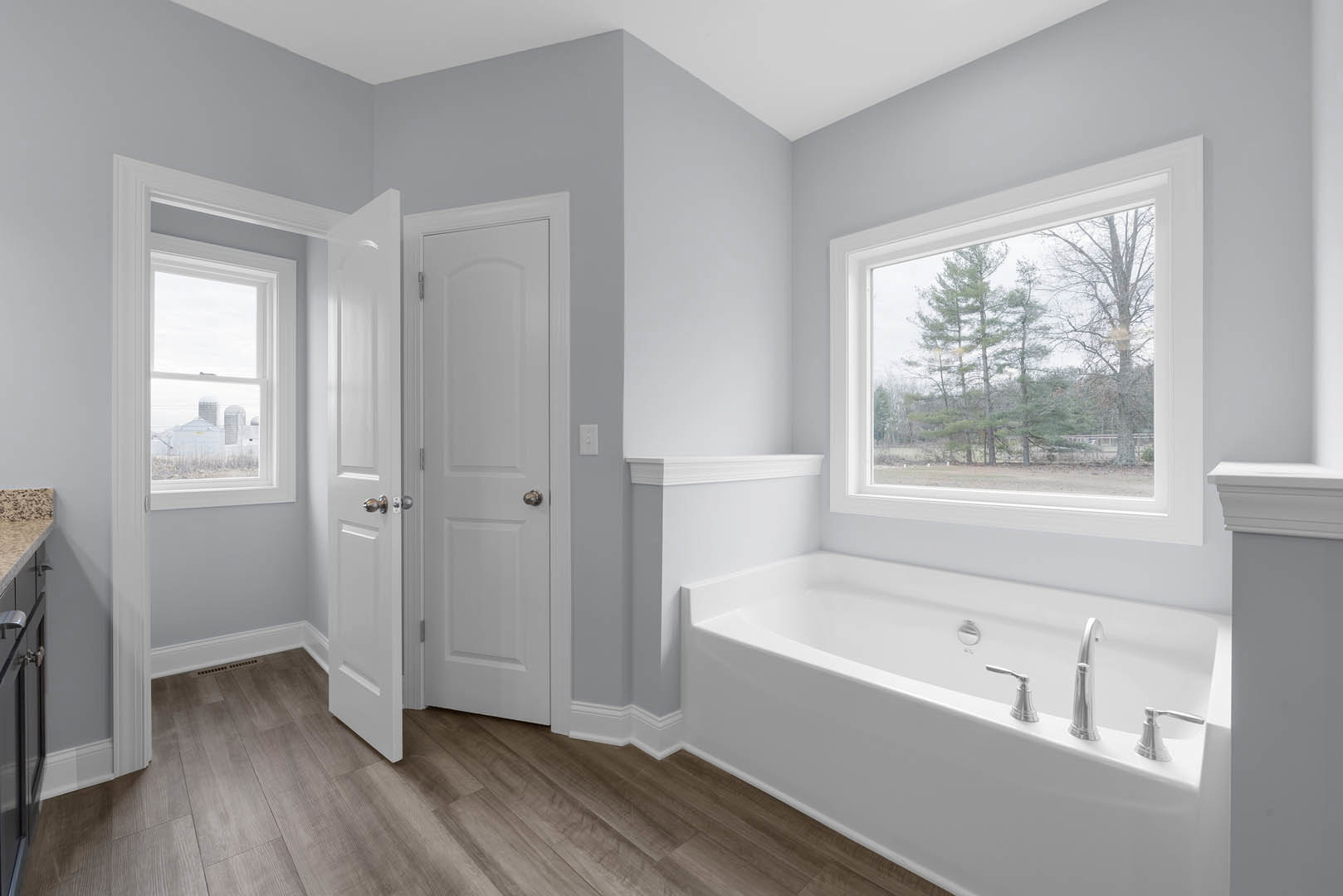Bathroom with freestanding bathtub, wood flooring, white door with silver doorknob, large window overlooking trees and farmland