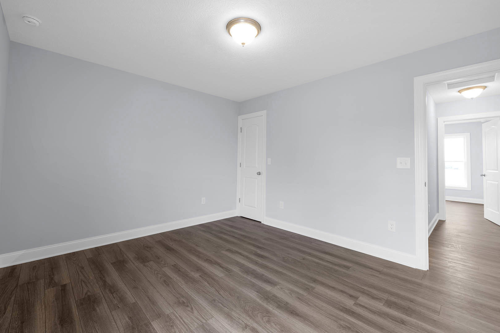White door with silver handle, wood laminate flooring, white-framed window, ceiling light fixture, plaster walls with crown molding