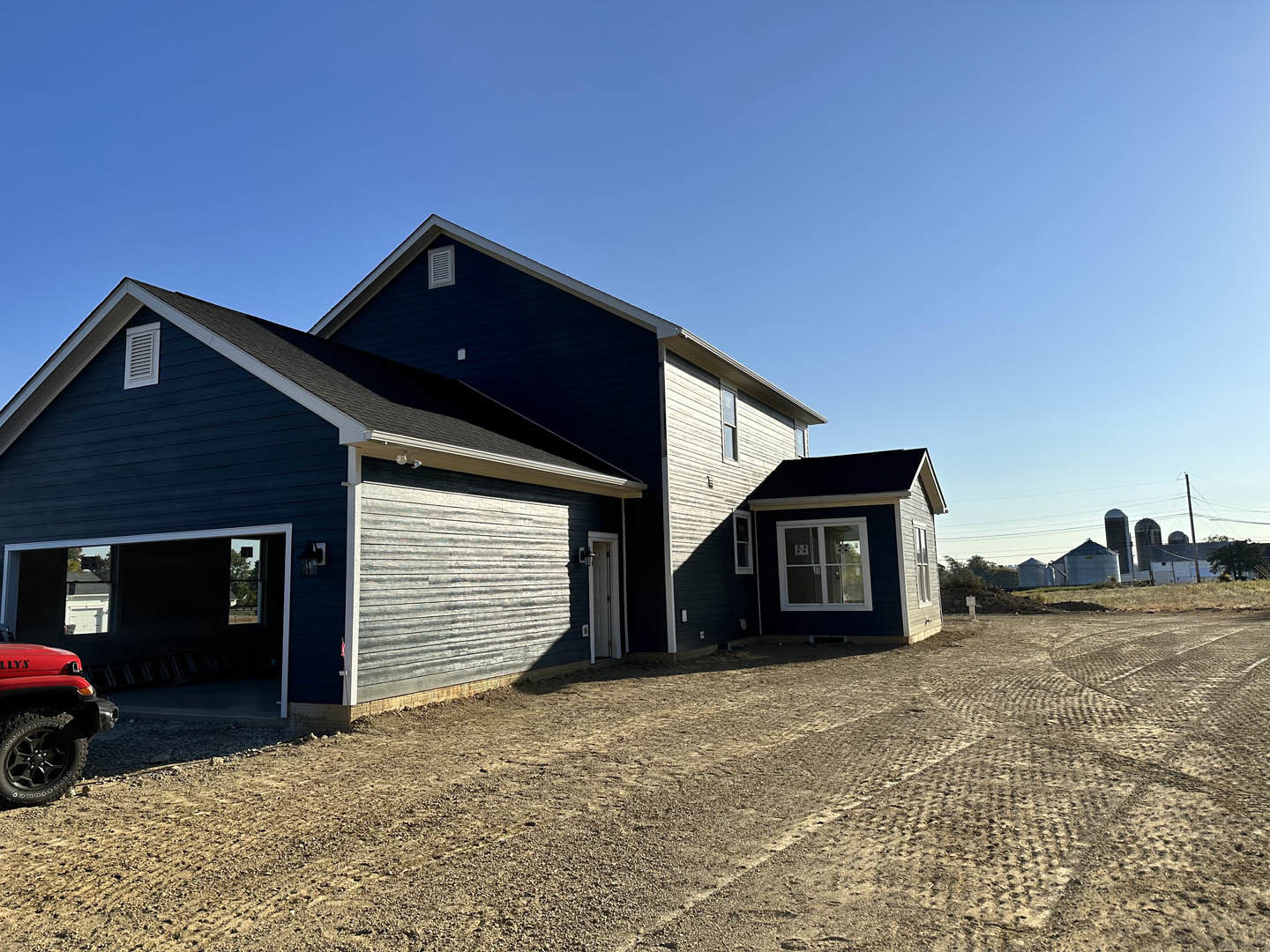 Partially built house with white-framed window, exposed wood siding, and blue sky, red jeep parked on dirt road in foreground
