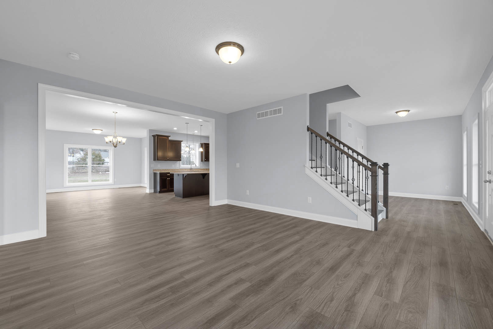 Open-concept room featuring wood flooring, white plaster walls, a modern staircase with metal railing, recessed ceiling lights, large window overlooking yard, and kitchen area with