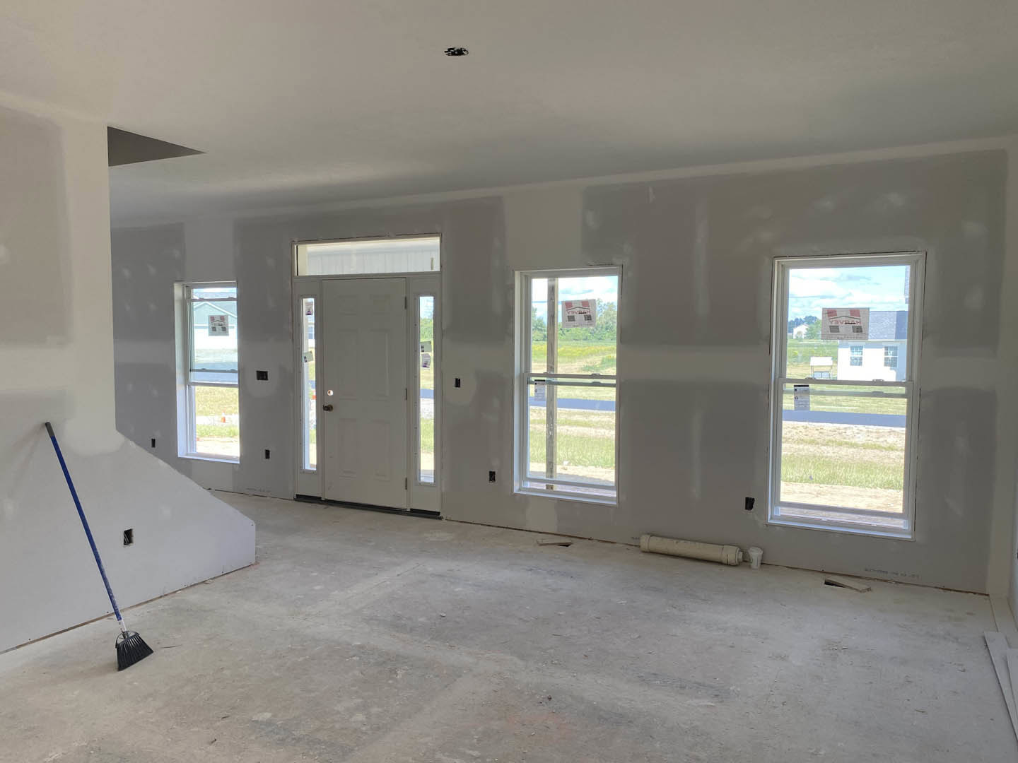 Bright room with smooth white plaster walls, large windows overlooking a grassy field, broom and blue stick leaning against wall, window features a sign with red and white text.