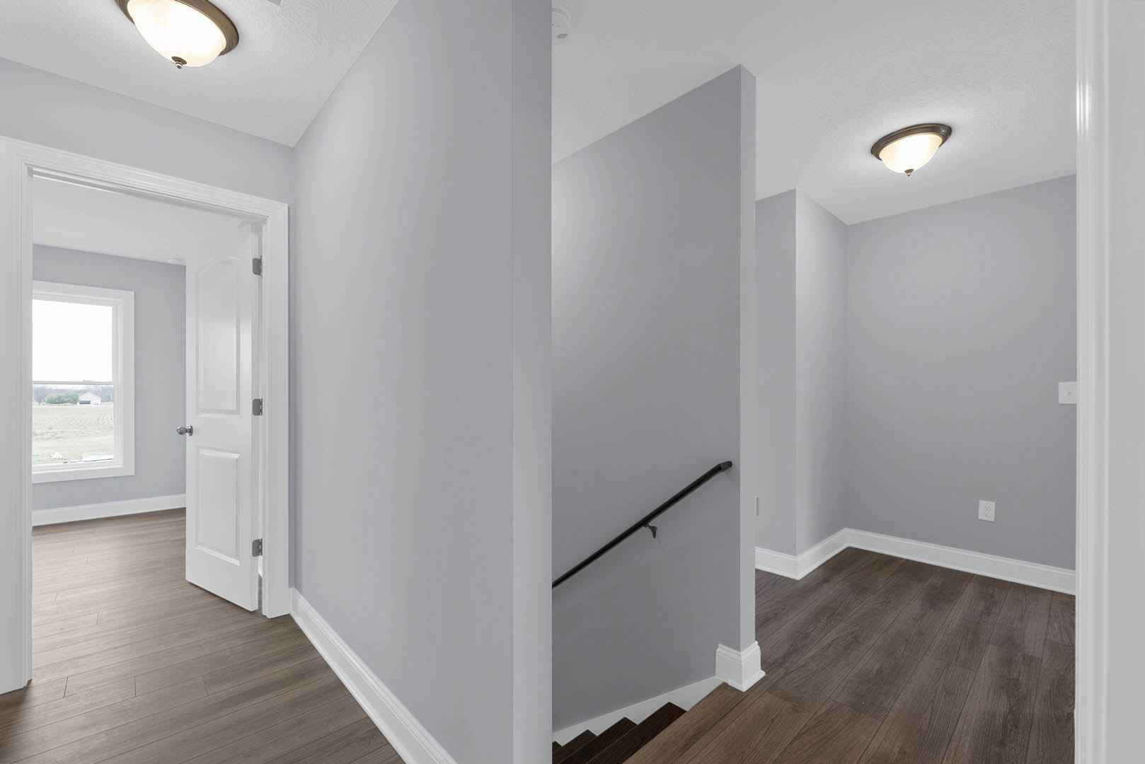 Hallway with wood laminate flooring, white plaster walls, staircase with wooden steps and railing, white door, ceiling light fixture, window overlooking grassy field.