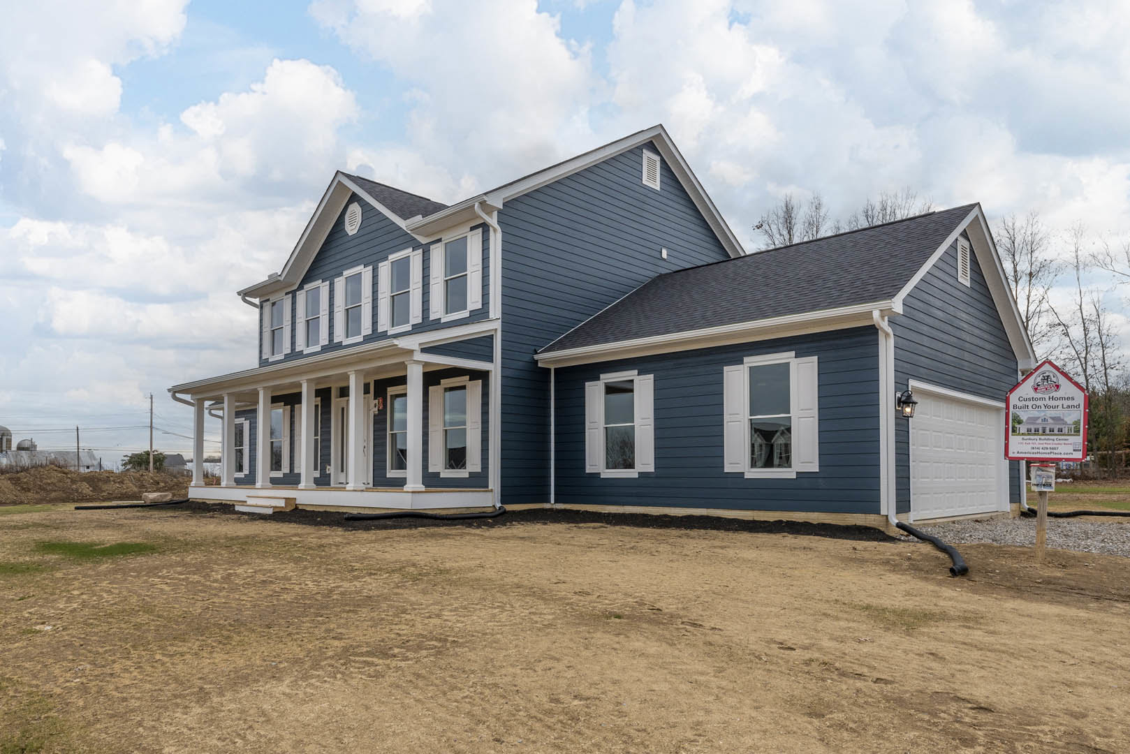Two-story house with blue siding and white trim, large grassy yard, covered porch, white-framed windows, real estate sign on post, cloudy sky overhead