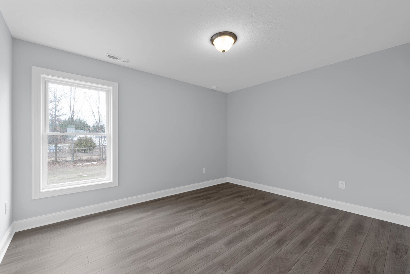 Sunlit room with wide window overlooking a grassy yard and distant trees, hardwood flooring, white plaster walls, ceiling light fixture, and decorative molding.
