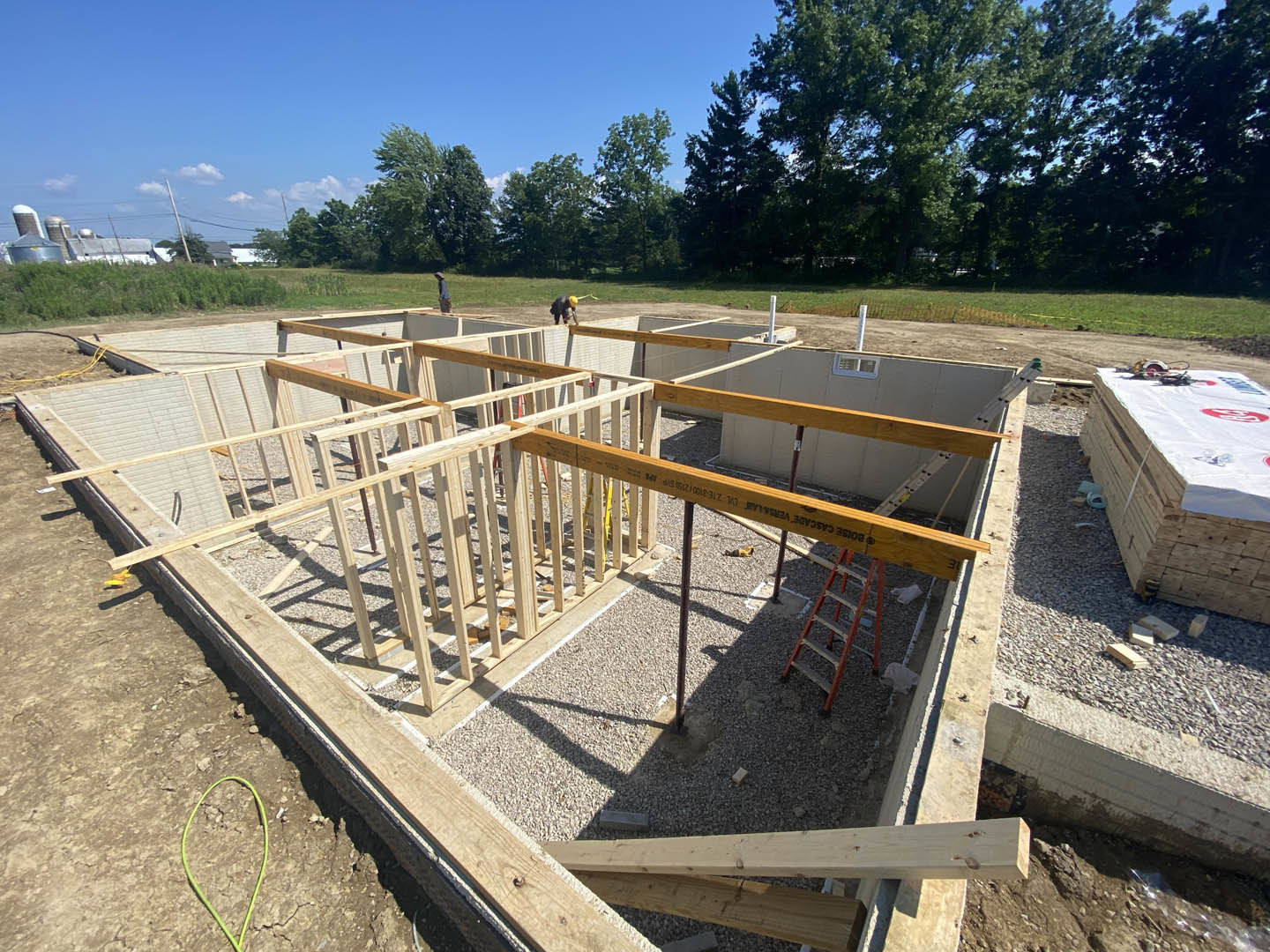 Partially built house foundation surrounded by stacks of wood planks, green wire on rocks, construction workers, and trees under a clear sky.
