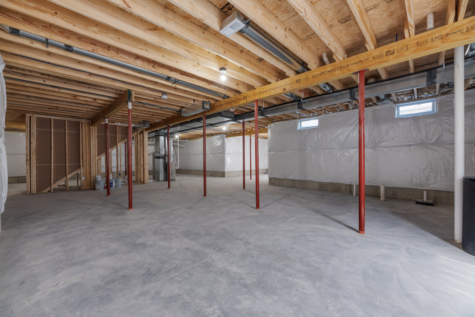 Basement room with exposed wooden ceiling beams, red structural poles, concrete and grey tile flooring, window wrapped in foil insulation, and visible metal pipe along the ceiling.