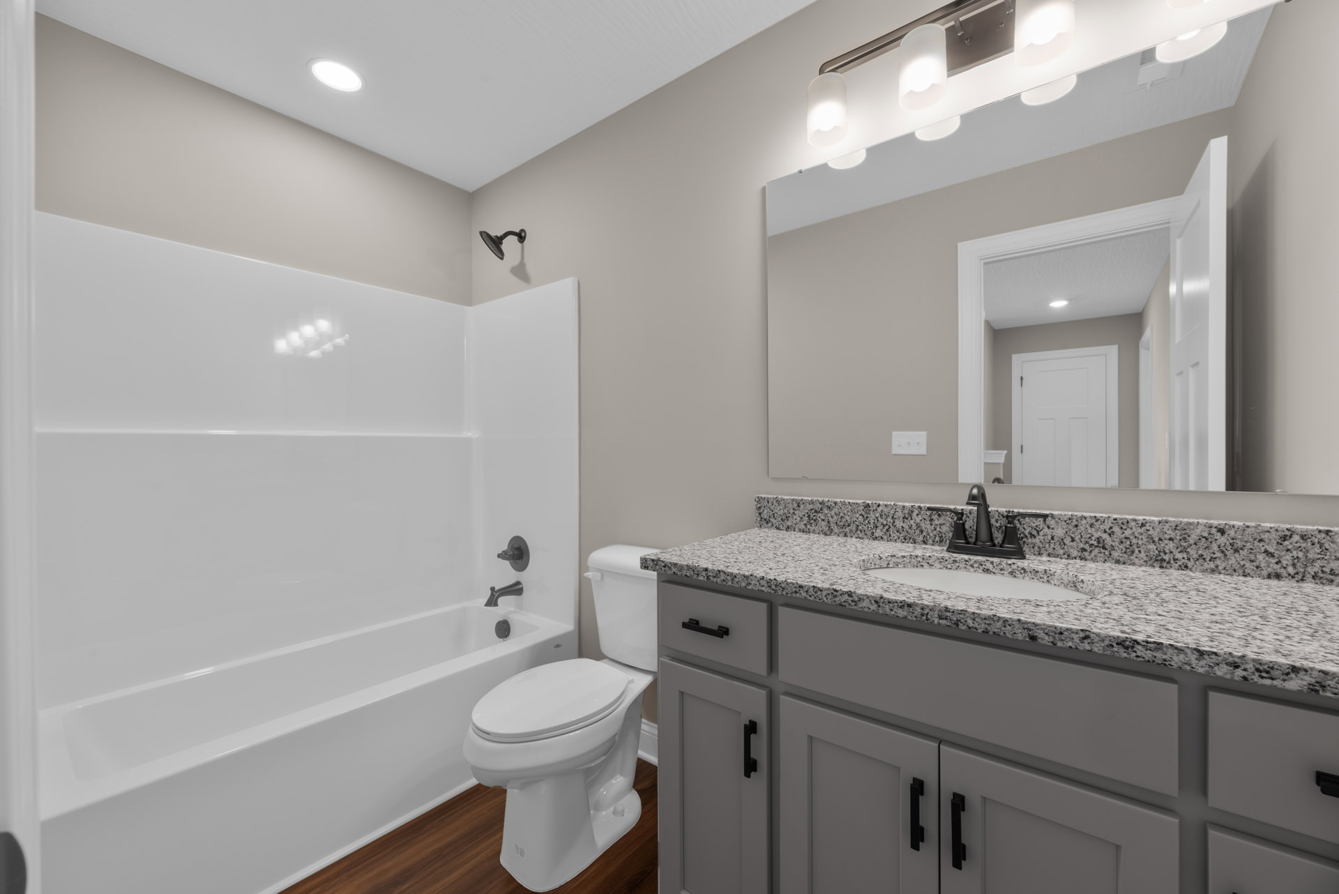 White bathtub with chrome shower head and faucet, adjacent to a white toilet; light gray tile walls, white door with black hinges, recessed ceiling light.