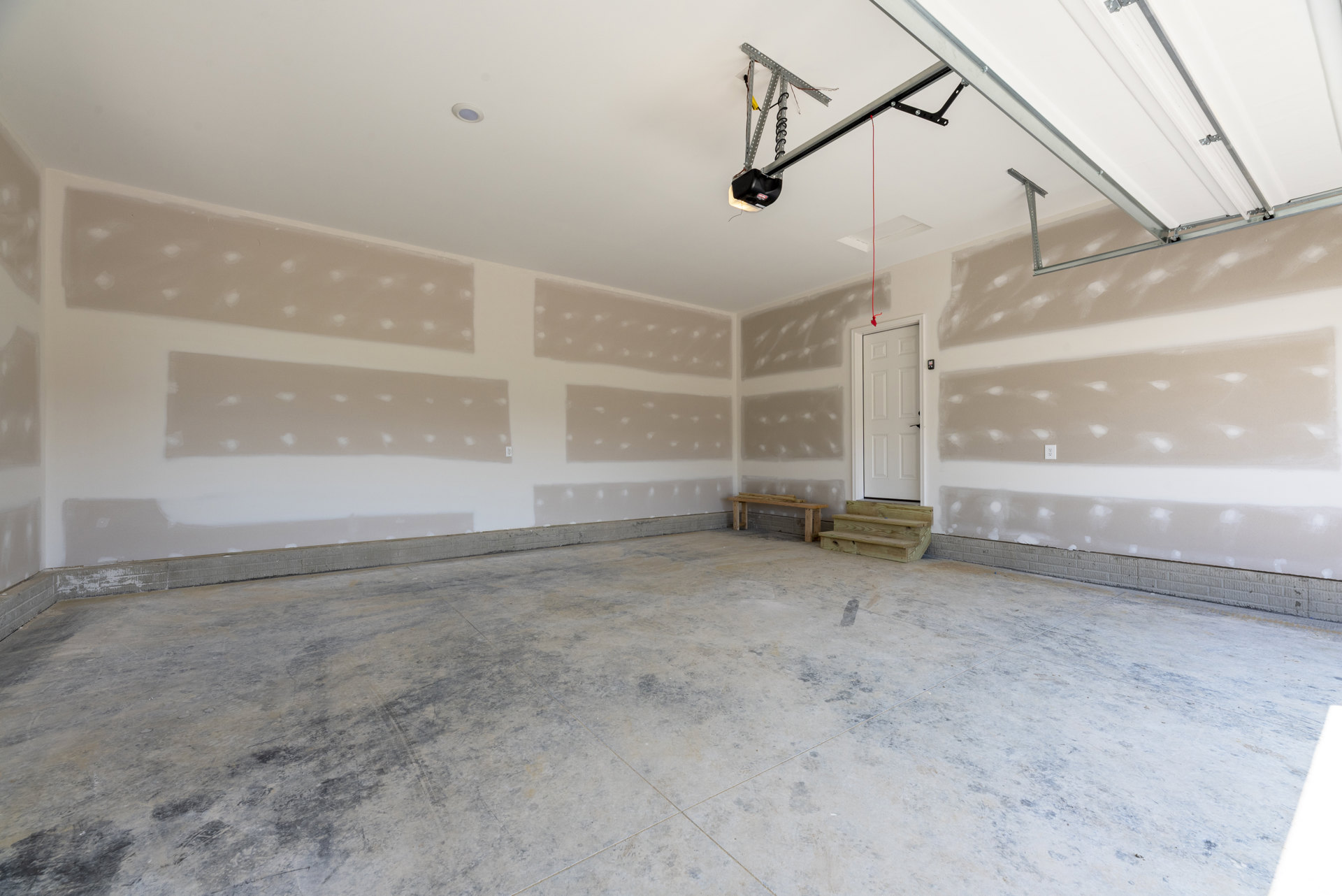 Concrete-floored room featuring a garage door, white plaster wall, wooden bench with wood plank seat, white door with black handle, and close-up of wooden staircase against gray