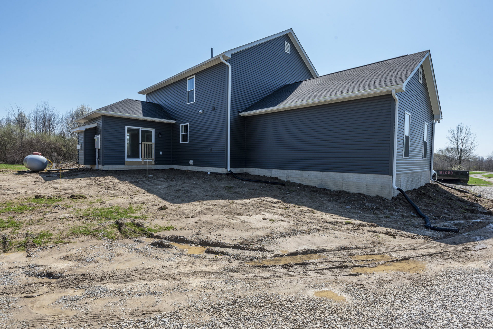 Single-story home with light siding, screen door and wooden railing, large grey water tank with red cap, dirt patch with tire tracks, black pipe on ground, sparse landscaping, tree