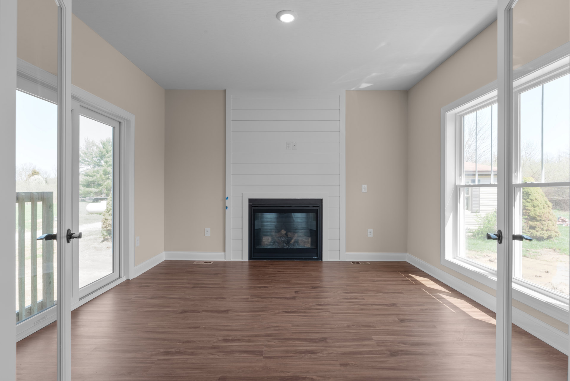 Living room with wood flooring, stone fireplace filled with logs, large windows, ceiling light fixture, and a vacuum cleaner near the window.