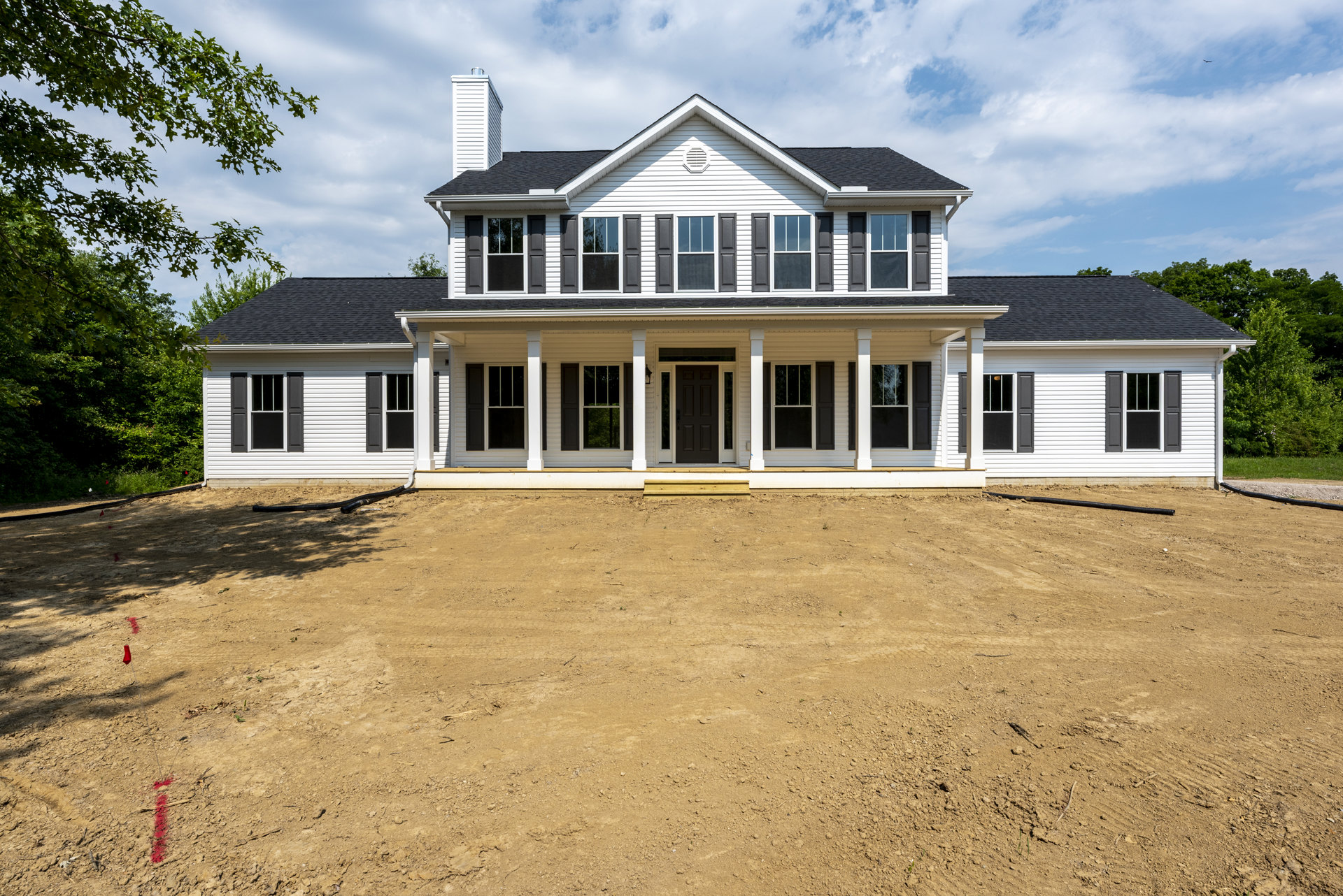 White two-story house with black front door, wide covered porch, multiple windows, and unfinished dirt yard under partly cloudy sky