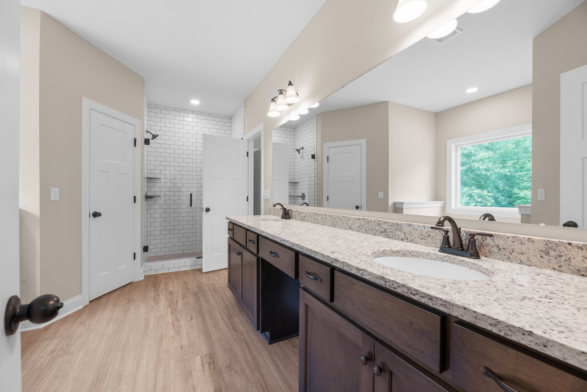 Bathroom with double sinks, wood flooring, white door with black handle, window showing trees, white countertops and cabinetry