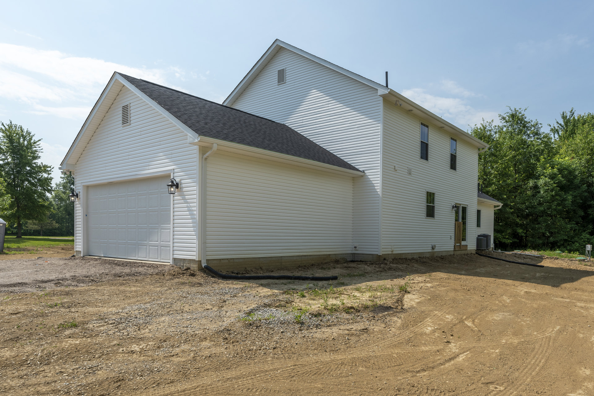 White siding house with attached garage, black pipe on dirt in foreground, leafy trees and cloudy sky in background