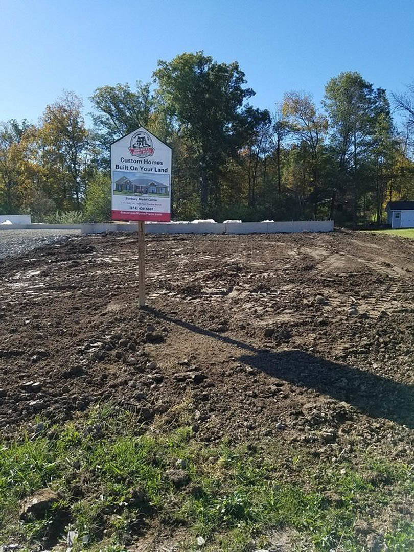Wooden post with a white sign featuring a black-roofed house illustration, standing in a bare dirt field with sparse grass and tree line in the background