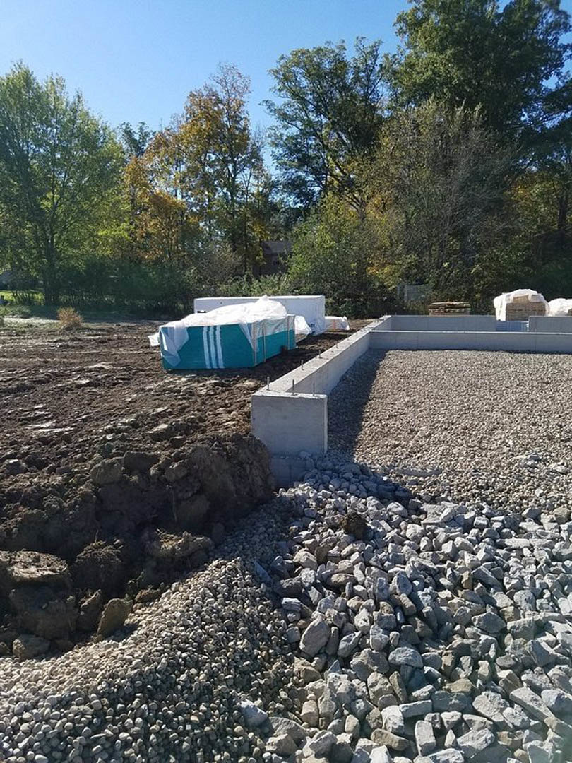 Gravel and concrete foundation surrounded by piles of rocks, blue and white cooler, leafy trees, and construction rubble on an outdoor site