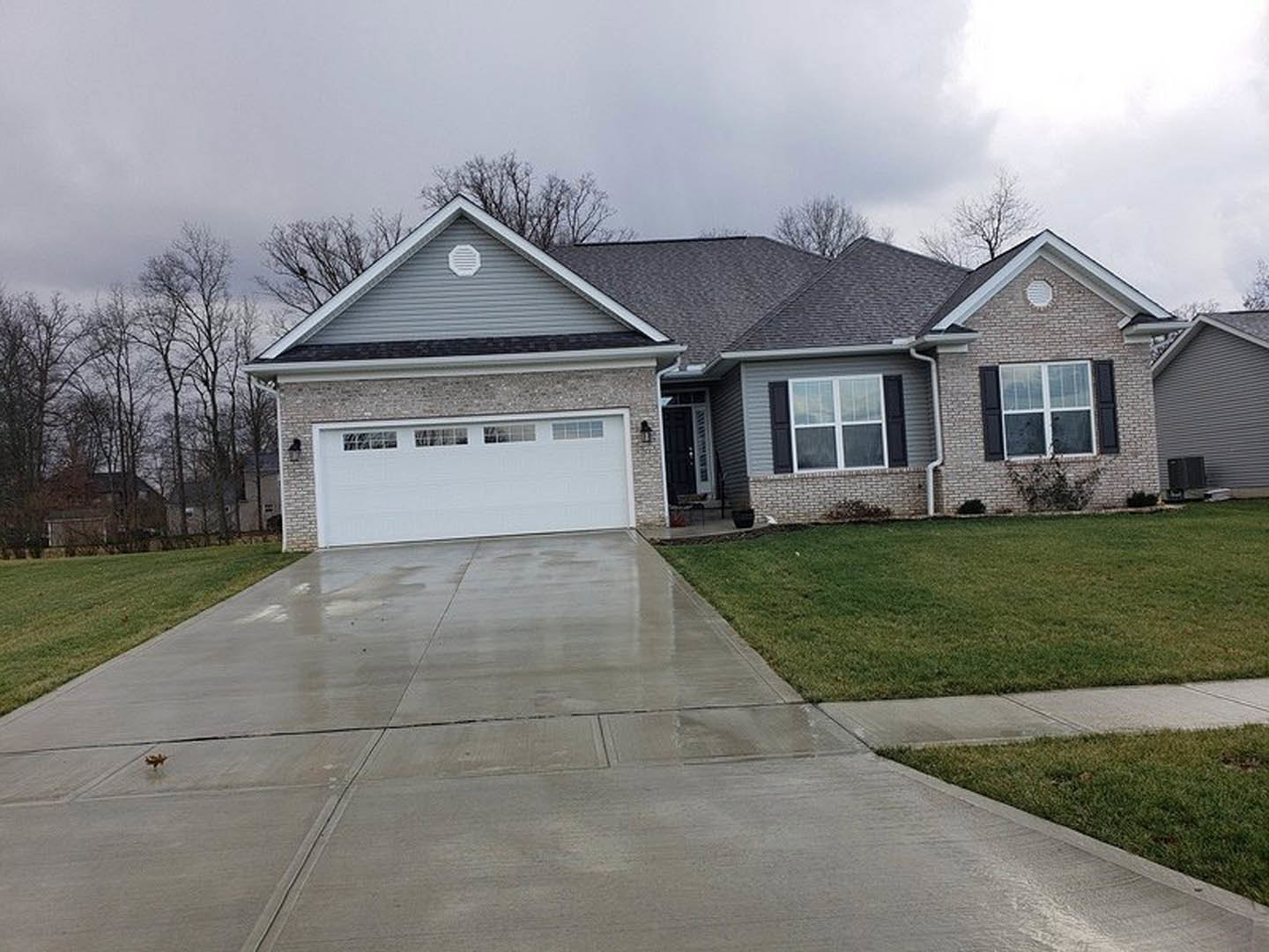 Modern home exterior with white siding, large windows, attached garage featuring windowed door, concrete driveway bordered by green lawn, landscaped shrubs, and mature trees under
