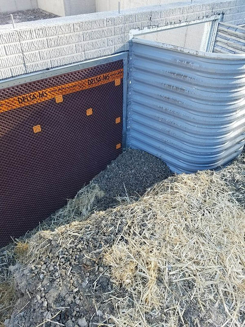 Metal drainage channel bordered by gravel and straw beside a white composite exterior wall