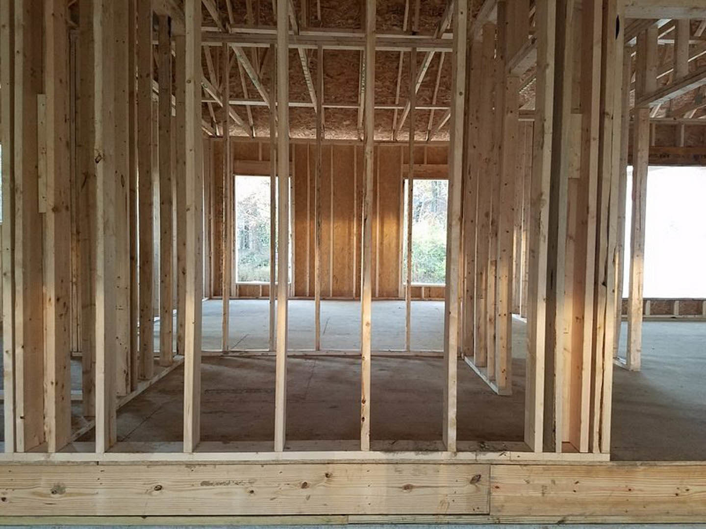 Room under construction with exposed wood framing, concrete floor, large windows, and visible wooden beams.