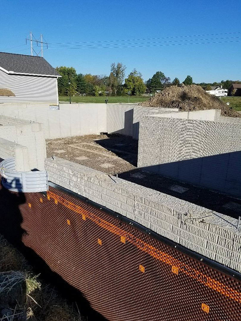 Construction site with large dirt piles, exposed concrete foundation, white plastic tubing, nearby green-leafed tree, and overhead power line under open sky.