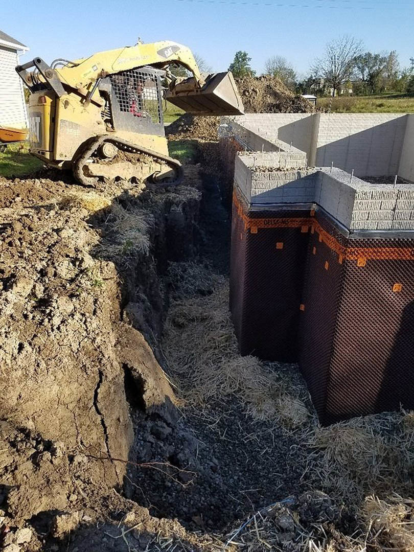 Bulldozer excavating soil on a residential construction site with exposed dirt, rocks, and partially built foundation, surrounded by trees and open sky