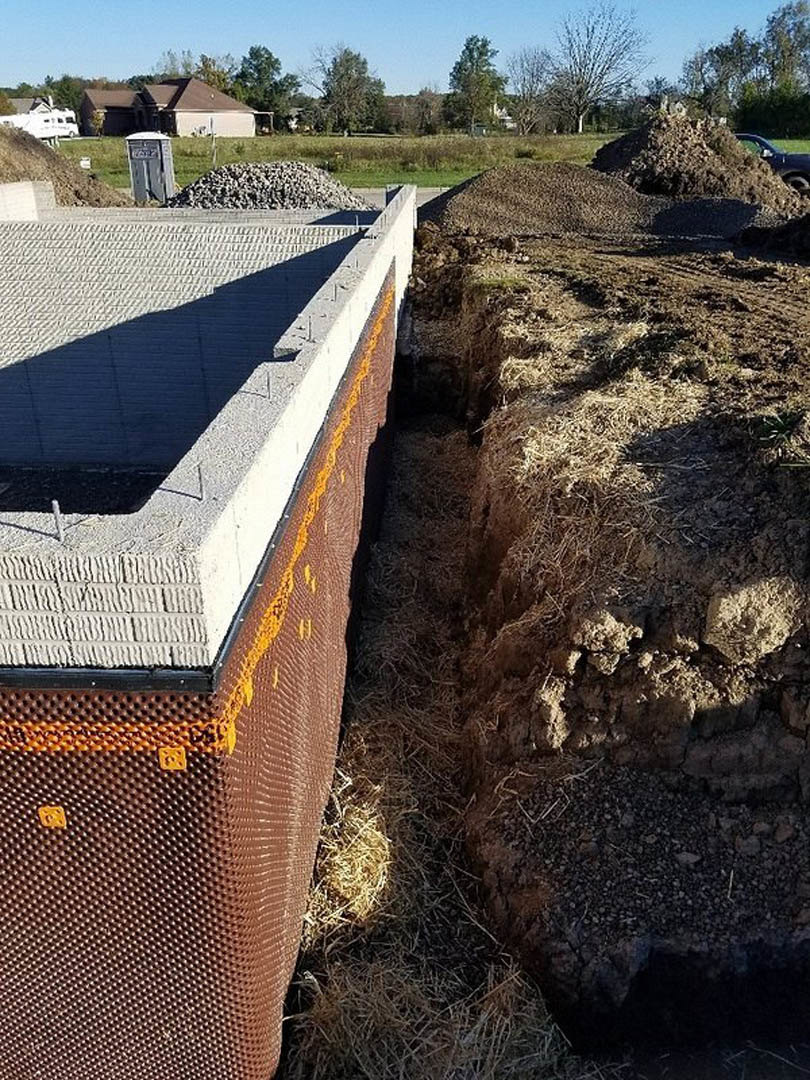 Concrete house foundation surrounded by dirt mounds, parked car, and construction materials; partially built house with roof visible in background, small white utility box with