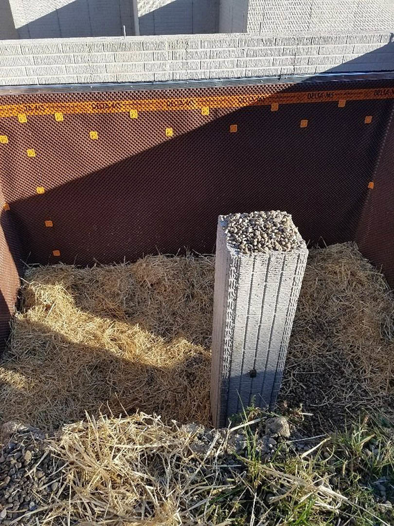 Concrete post set in a grassy fenced yard, surrounded by a white fence, brick wall, and piles of rocks and straw.