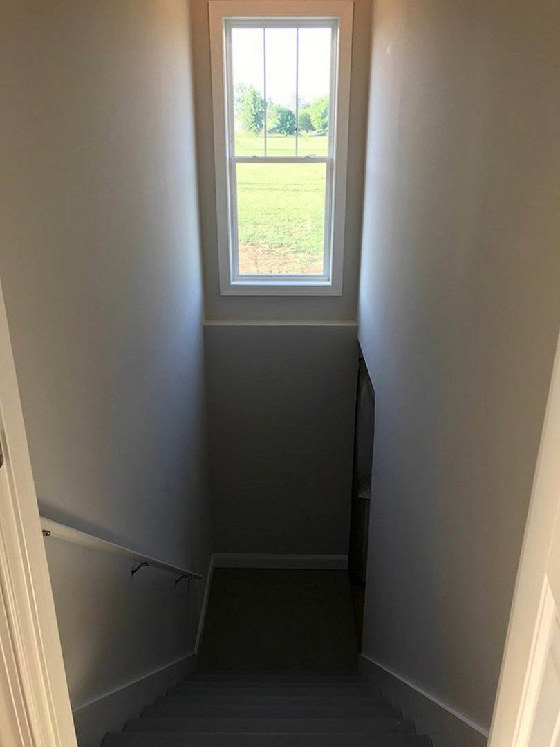 Hallway with plaster walls, dark flooring, and a large window overlooking grassy yard; stairs visible in shadowed area.