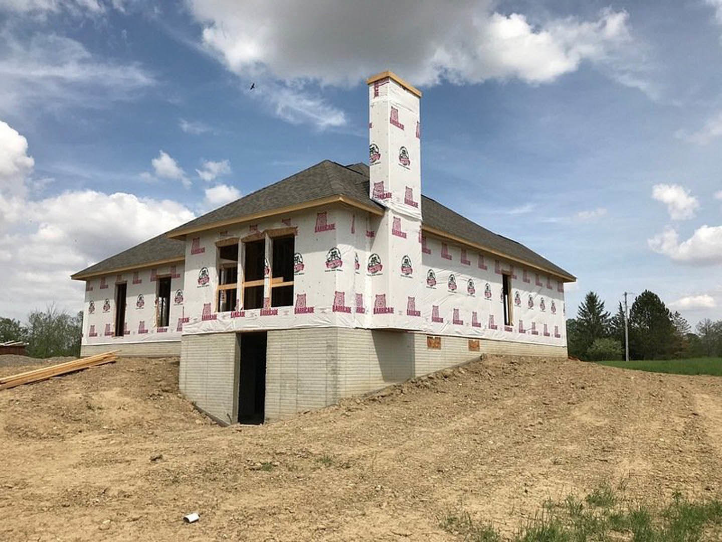 Partially built house with white chimney, exposed framing, dirt lot in foreground, grassy area, and cloudy sky in background