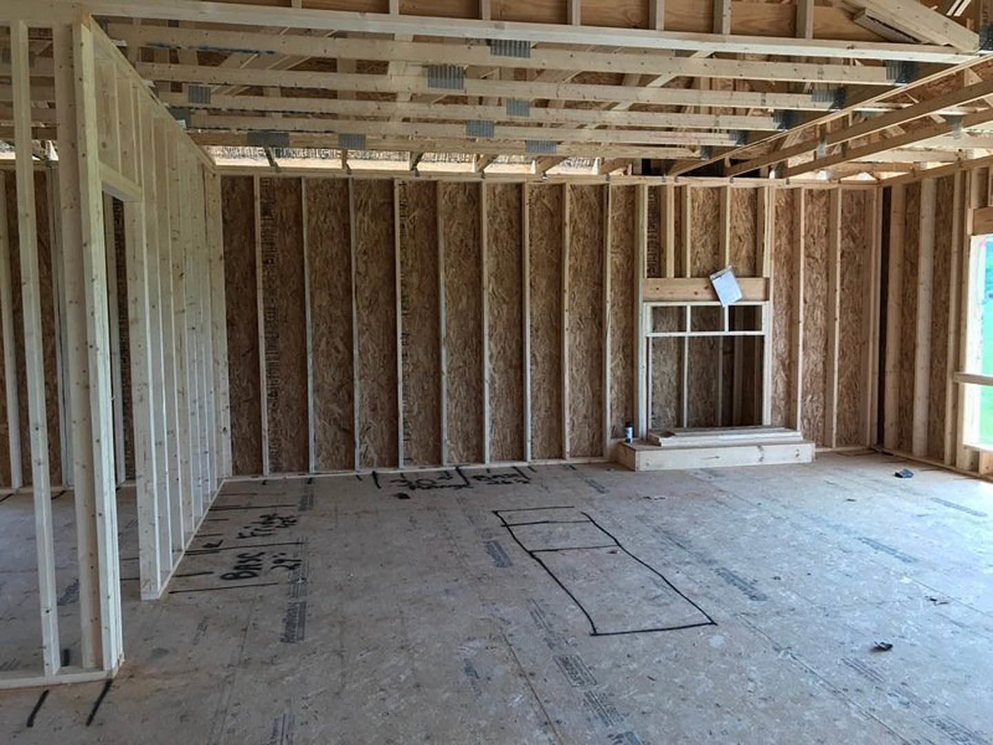 Open room with exposed wood ceiling beams, unfinished floor marked with construction drawings, large window framed in wood, clipboard resting on windowsill, white rectangular