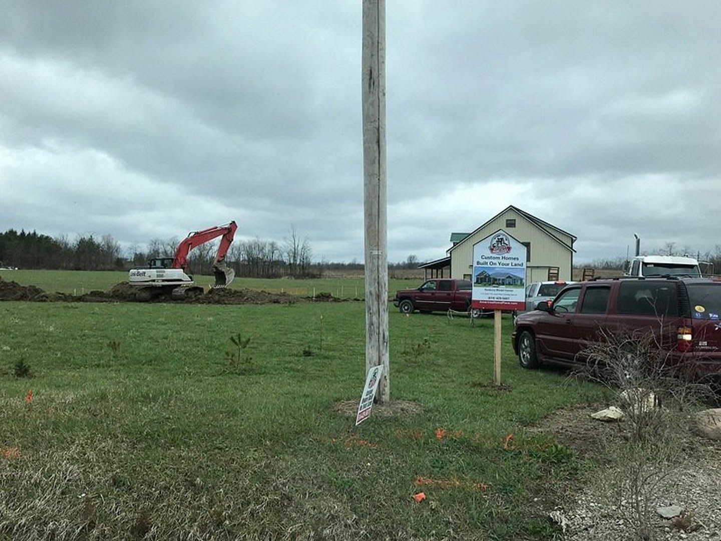 Framed custom home under construction with crane, red excavator digging foundation, trucks and cars parked nearby, grassy lot, wooden pole with house sign, cloudy sky overhead