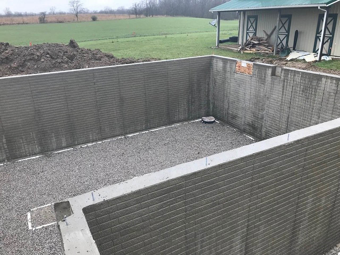 Concrete retaining wall bordered by gravel, wooden planks stacked in front of a modern house, grassy ground, and trees in the background
