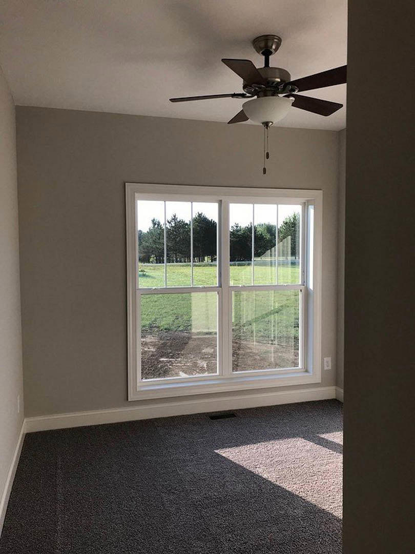 Ceiling fan with light fixture above carpeted floor, large window revealing grassy field and trees, neutral walls and bright daylight filling custom home room