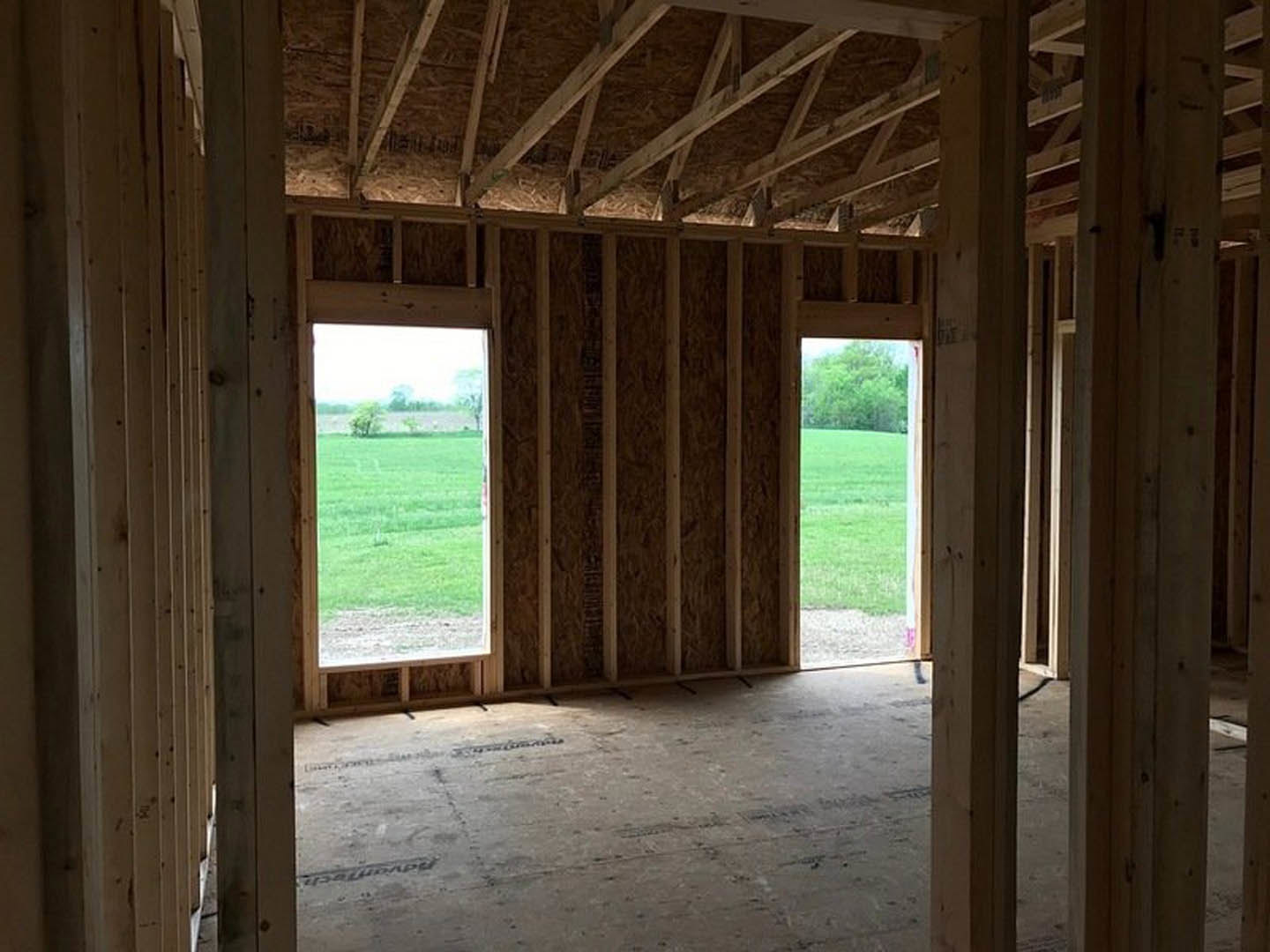 Open room with exposed wooden roof trusses, concrete floor, large window overlooking grassy field and open landscape