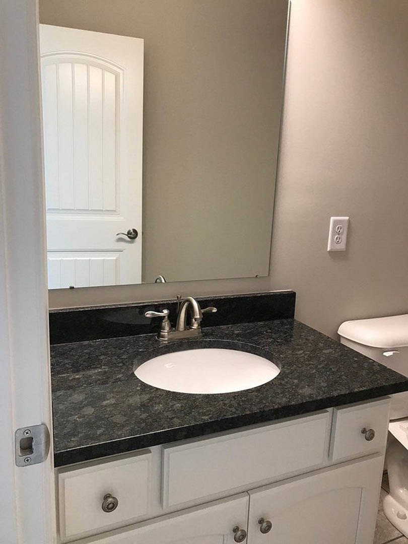 Bathroom with black stone countertop, rectangular mirror, white plate, chrome faucet, white cabinetry, and tile wall.