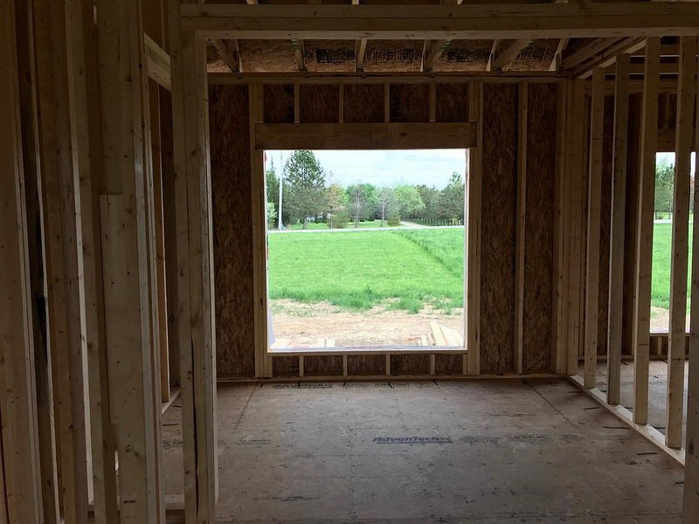 Large window framed with natural wood beams overlooks a grassy field and cluster of trees, concrete floor visible inside the room.