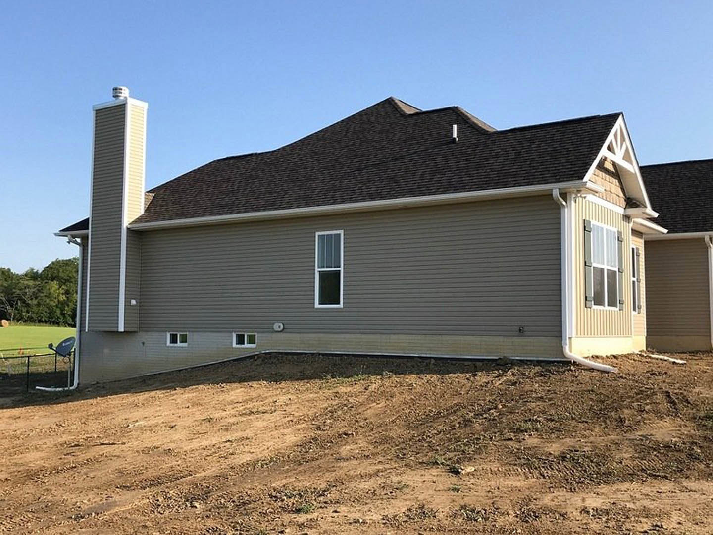 White-sided house with gray roof, side chimney, and white-framed window; dirt hill and additional white building in background under blue sky