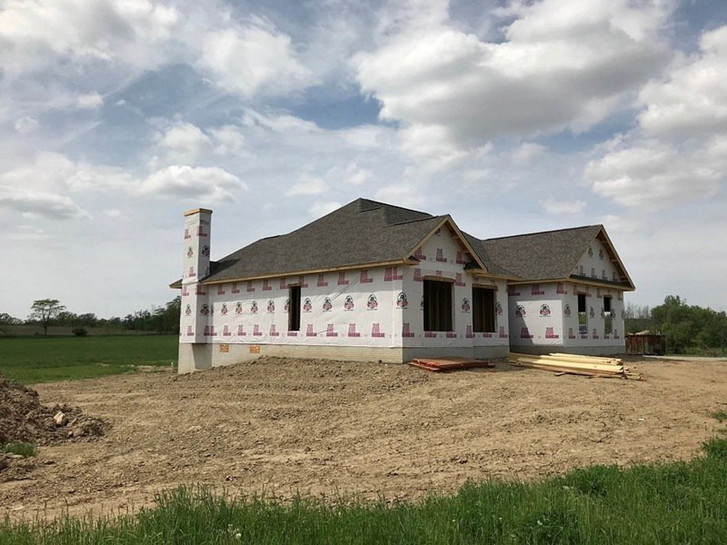 Partially built house with shingled roof, exposed framing, dirt lot in foreground, patches of grass, distant tree, and chimney visible against cloudy sky
