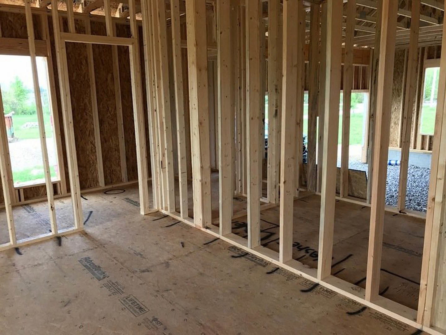 Exposed wood framing and beams in an unfinished room, window opening overlooking grassy yard, plywood subfloor visible