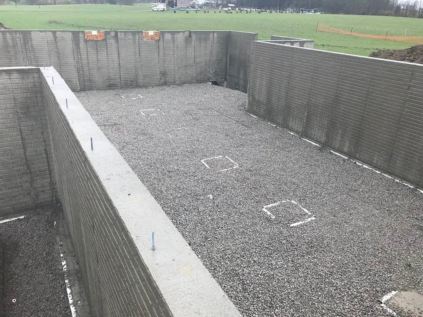 Concrete exterior wall featuring a geometric square pattern, white painted line on gravel ground, sparse grass and plants along fence.